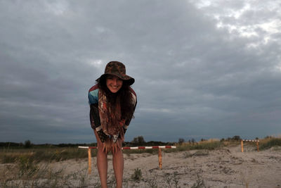 Portrait of smiling young woman standing on field against cloudy sky during sunset