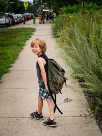 Side view of smiling boy with backpack walking on footpath amidst plants in park