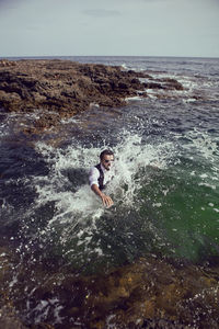 Man with a beard and sunglasses in clothes a vest and a white shirt jump in the sea among the rock