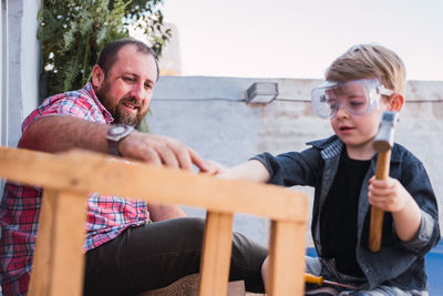 Side view of hipster dad helping son on protective glasses with hammer while doing woodwork