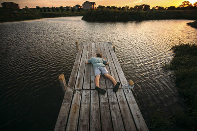 High angle view of boy lying down on jetty against sky during sunset