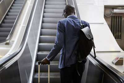 Rear view of businessman standing with luggage moving up on escalator at subway station