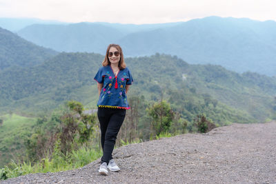 Adult traveling woman wearing sunglasses standing and smiling on the top of the mountain cliff 