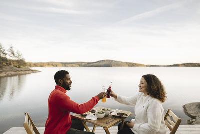 Smiling couple toasting bottles while sitting on chair by lake during weekend