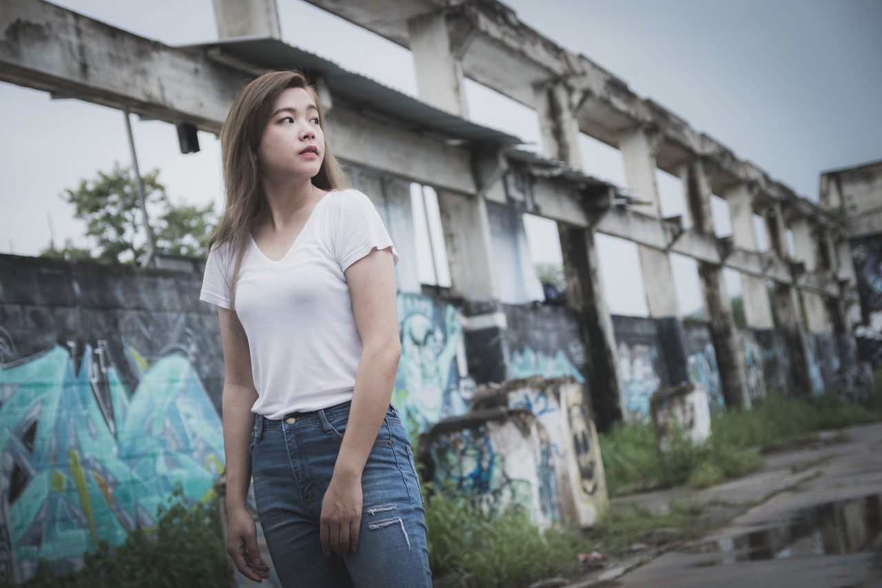 BEAUTIFUL YOUNG WOMAN LOOKING AWAY WHILE STANDING AGAINST BUILT STRUCTURE