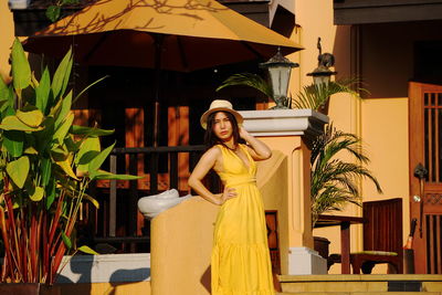 Portrait of smiling woman wearing dress and hat standing against house