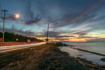 Road by illuminated street against sky during sunset