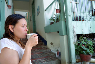 Side view of thoughtful woman having drink while standing against house