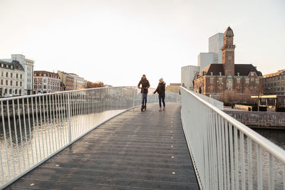 Teenage girl walking with friend riding push scooter on bridge in city against clear sky