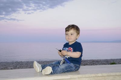 Cute toddler boy sitting with car key on retaining wall while looking away at beach against sky