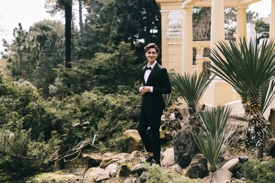 A beautiful young man, the groom in an elegant wedding suit, stands posing in the city's old park