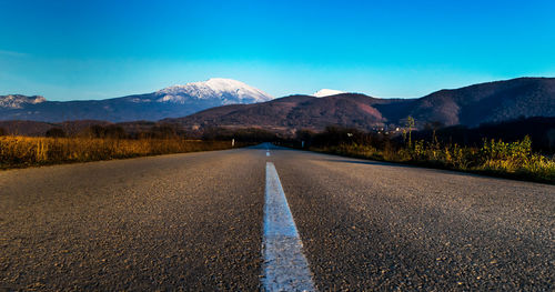 Empty road by mountains against clear blue sky