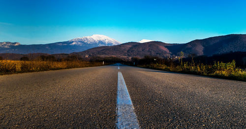 Empty road by mountains against clear blue sky