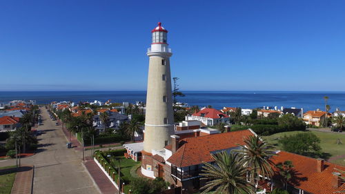 Lighthouse amidst buildings and sea against clear blue sky