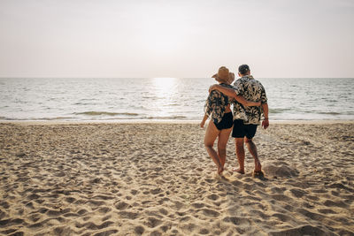 Carefree senior couple walking with arms around on sand at beach