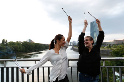 Cheerful young friends holding sparklers while standing against river in city