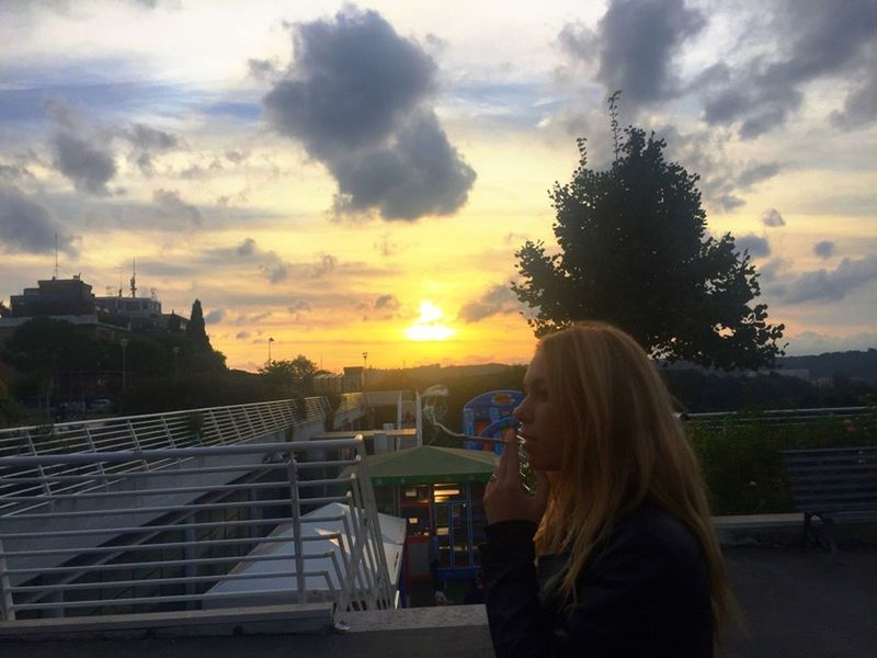 sunset, sky, lifestyles, leisure activity, rear view, cloud - sky, person, waist up, sitting, orange color, building exterior, casual clothing, cloud, sun, transportation, long hair, built structure, standing