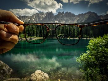 Cropped hand holding eyeglasses against lake and rocky mountains