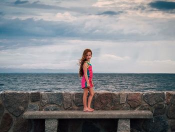 Portrait of girl standing on park bench in front of sea against cloudy sky