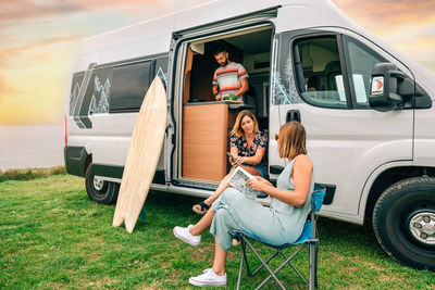 Two women talking in front of camper van while young man cooks