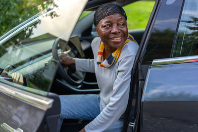 Side view of african woman sitting in electric car with open door smiles backward.