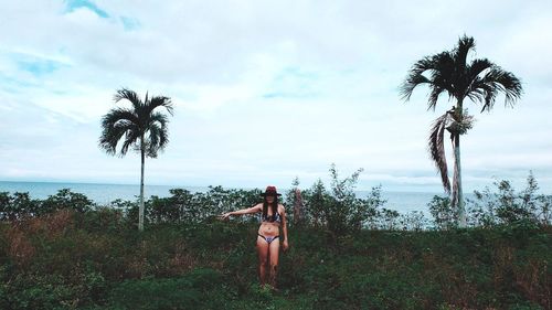 Portrait of young woman in bikini standing on grassy field by sea against cloudy sky