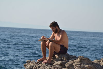 Young man sitting on rock by sea against sky