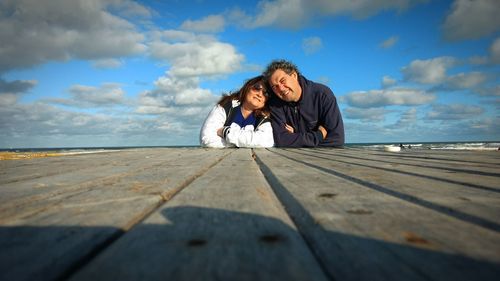 Couple leaning on boardwalk at beach against cloudy sky