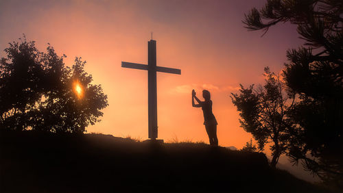 Silhouette cross standing by trees against sky during sunset