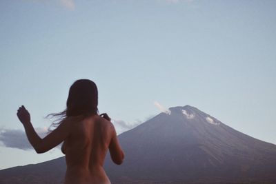 Rear view of naked woman standing by mountain against clear sky