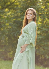 Young pregnant woman stands in park,looks at camera, green trees on background. brown haired female