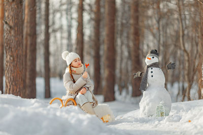 Smiling teenage girl sitting by snowman outdoors