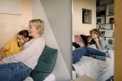 Smiling boy lying on bed while looking at mother and sister seen through mirror