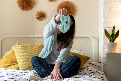 Middle aged woman in pajamas and sleep mask sitting relaxed on bed at home