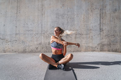 Slim female athlete with eyes closed in sportswear sitting on mat and stretching arms during workout in city