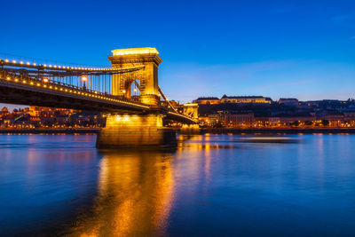 Illuminated bridge over river against blue sky in city