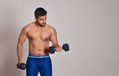 Shirtless muscular young man holding dumbbells against wall