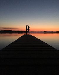 Mid distance view of silhouette couple standing on pier at lake against clear sky during sunset