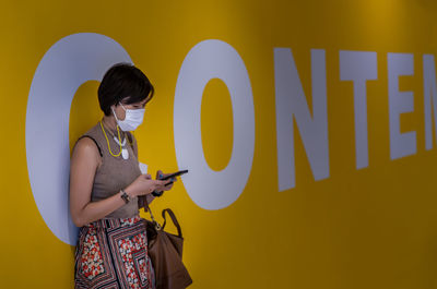 Woman wearing mask using mobile phone while standing by wall