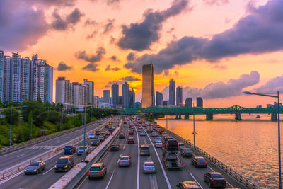 Cars on road by buildings against sky during sunset