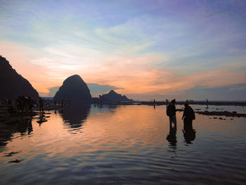 People wading in sea against sky during sunset