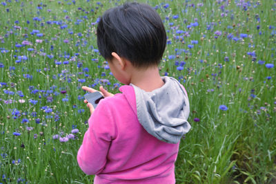 Rear view of girl holding smartphone against wild flowers on field