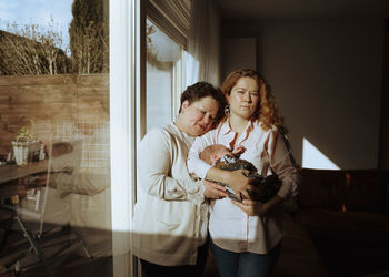 Portrait of young woman holding her baby son and  standing together with mother at home next to wi