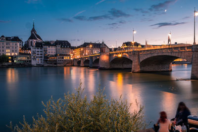 Illuminated bridge over river against buildings at dusk