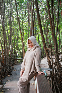 Portrait of smiling woman in hijab while standing on footbridge in forest