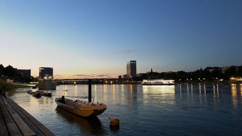 Boats in river by illuminated buildings against sky during sunset