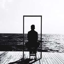 Rear view of silhouette man sitting on pier over sea during sunny day