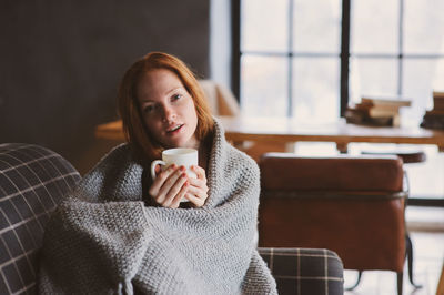 Portrait of young woman wrapped in blanket holding coffee cup on sofa at home