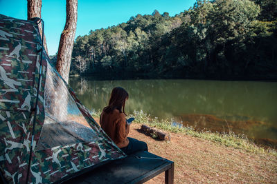 Rear view of woman sitting by lake against trees