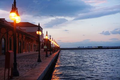 Illuminated building by sea against sky at sunset